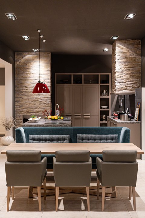 Moderner Essbereich mit Holztisch, blaugrüner Bank und grauen Stühlen, angrenzend an eine Küche mit hellbraunen Schränken und Steinelementen
